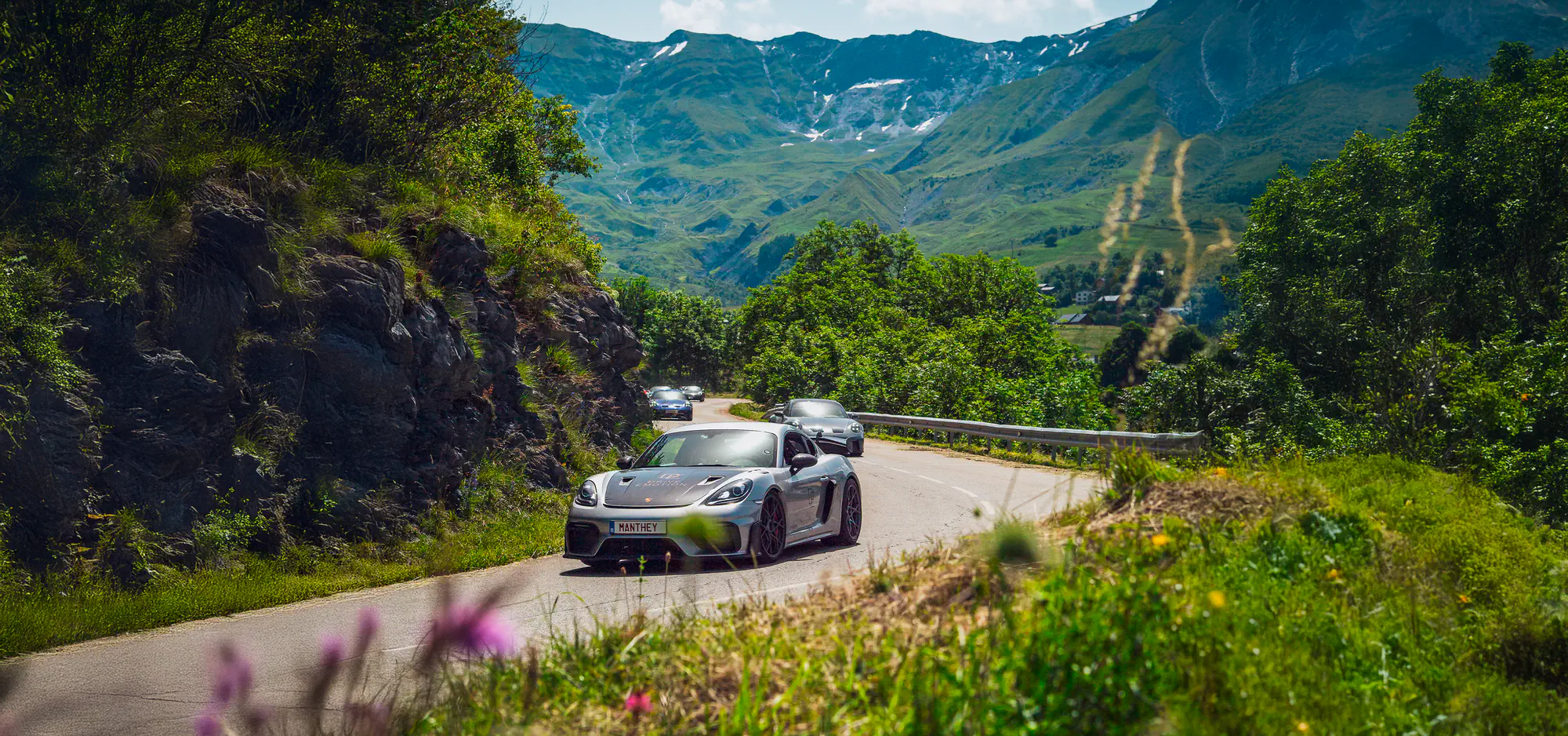 Porsche grise dans un virage d’une route de montagne bordée de fleurs, road trip Évasion Alpine