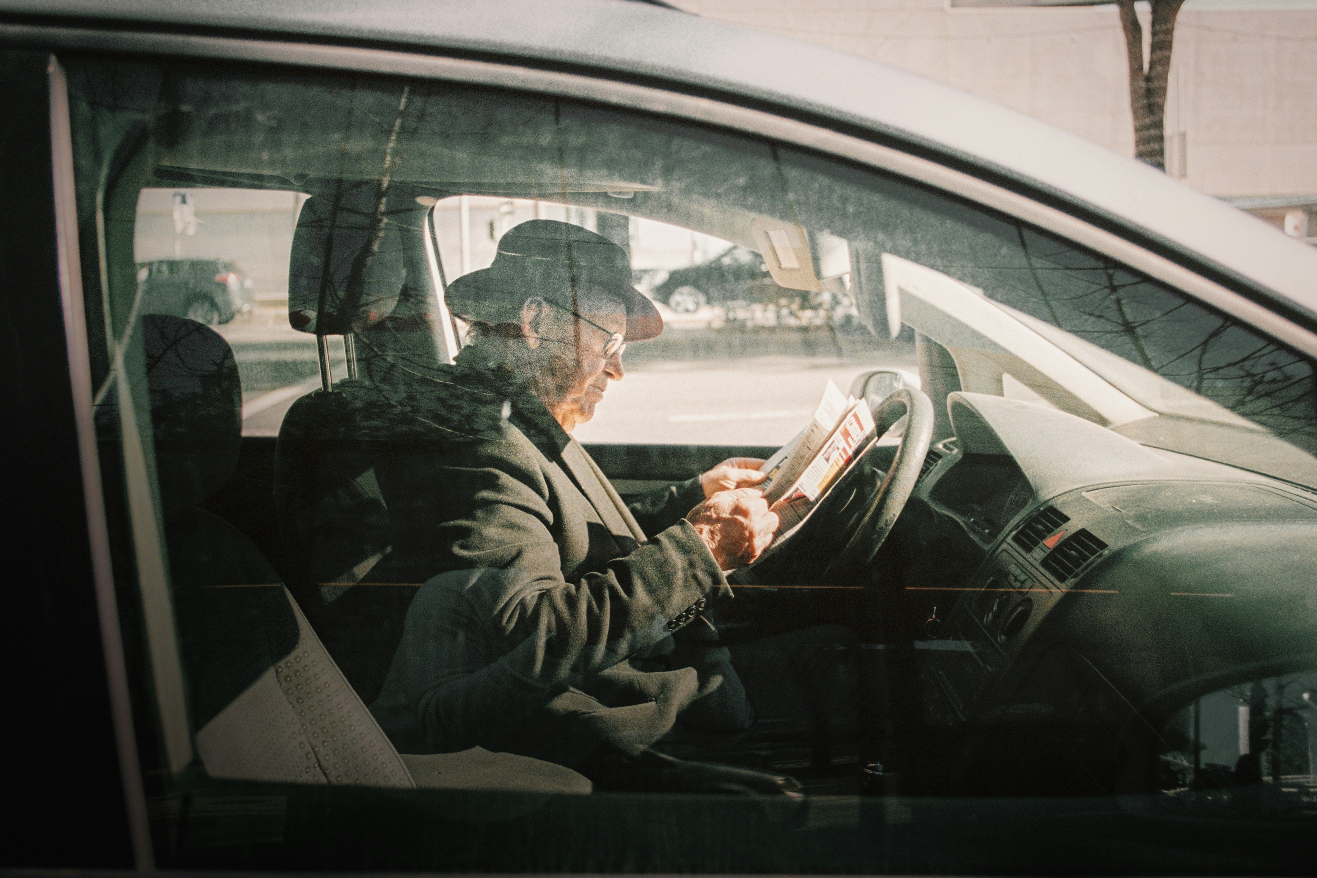 An older man reading a newspaper in the driver's seat of a car 