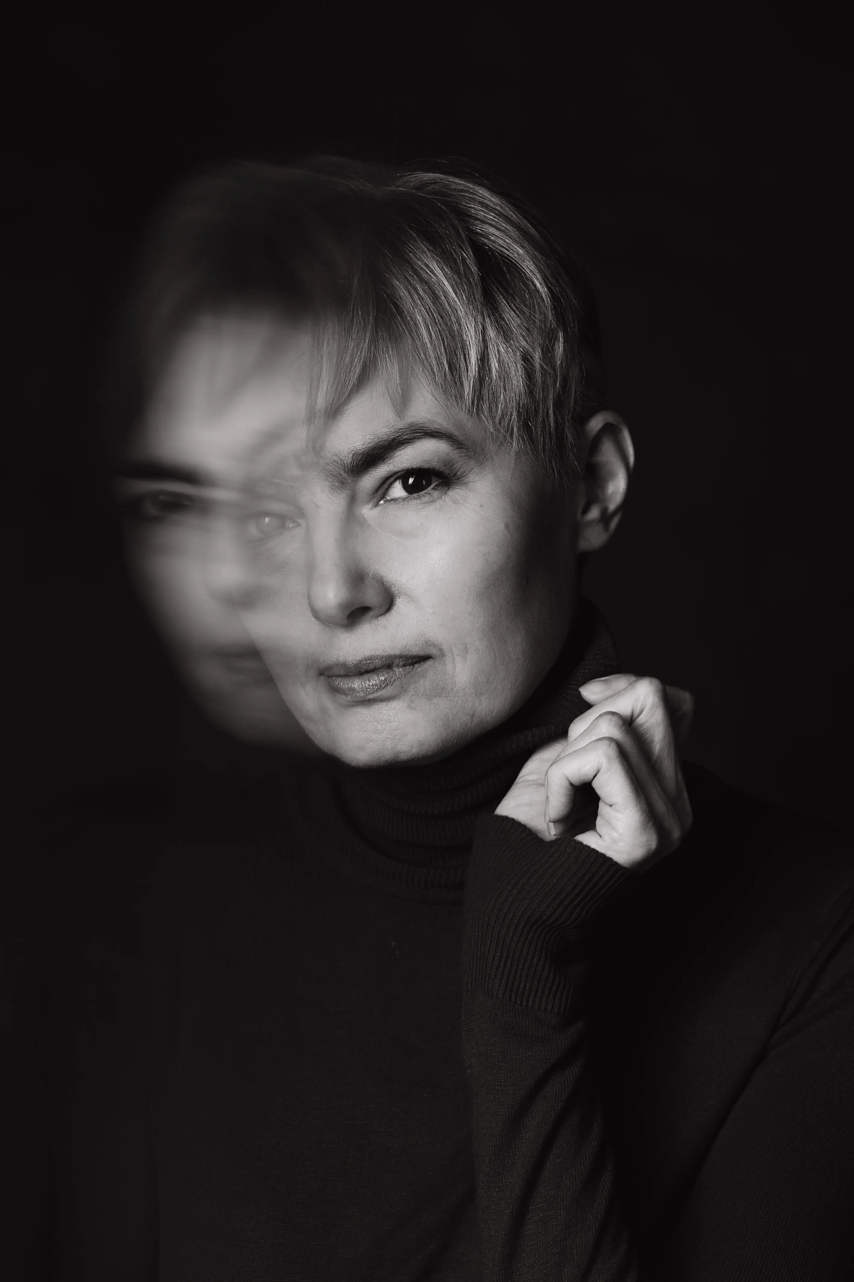 Czarno-biały portret kobiety o krótkich włosach pozującej do zdjęcia. Część twarzy jest rozmazanym, lustrzanym odbiciem.
