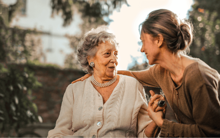 Caregiver and elderly woman smiling and holding hands outdoors.