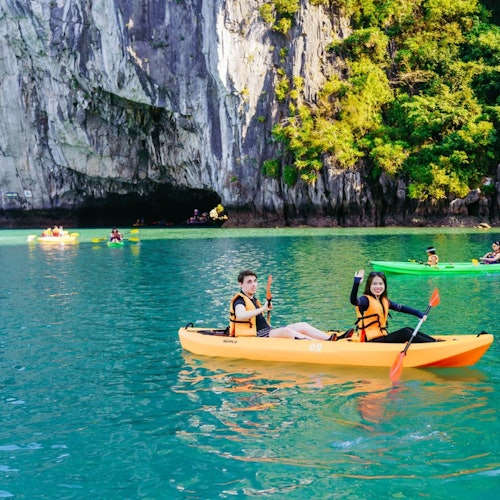 To personer i en gul kajak padler på turkis vand nær en klippefyldt klippe og grønne omgivelser, med andre kajakroere i baggrunden.