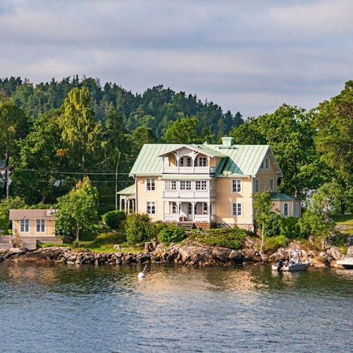 Gult hus med grönt tak vid vattnet, omgivet av träd. En liten brygga och båt syns till höger.
