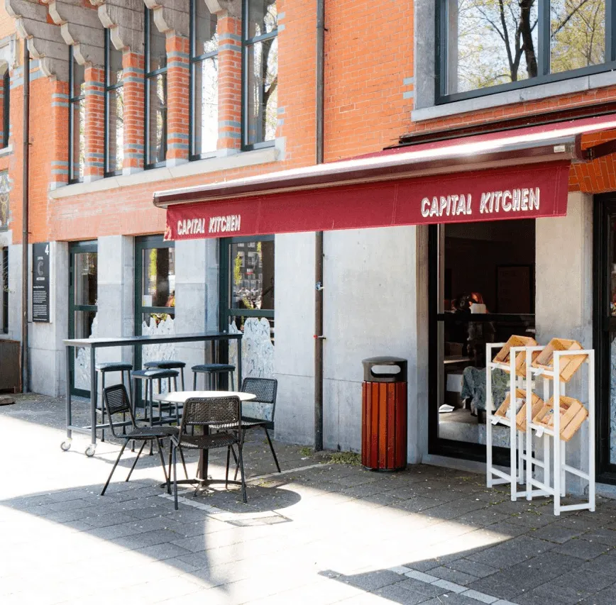 Outdoor view of a café with red awnings, tables, and chairs in a sunny urban setting.