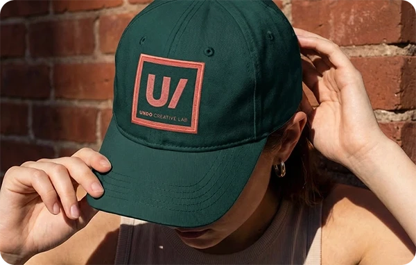 Close-up of woman wearing dark green Undo Creative Lab branded dad cap with coral logo patch, adjusting brim against sunlit brick wall background