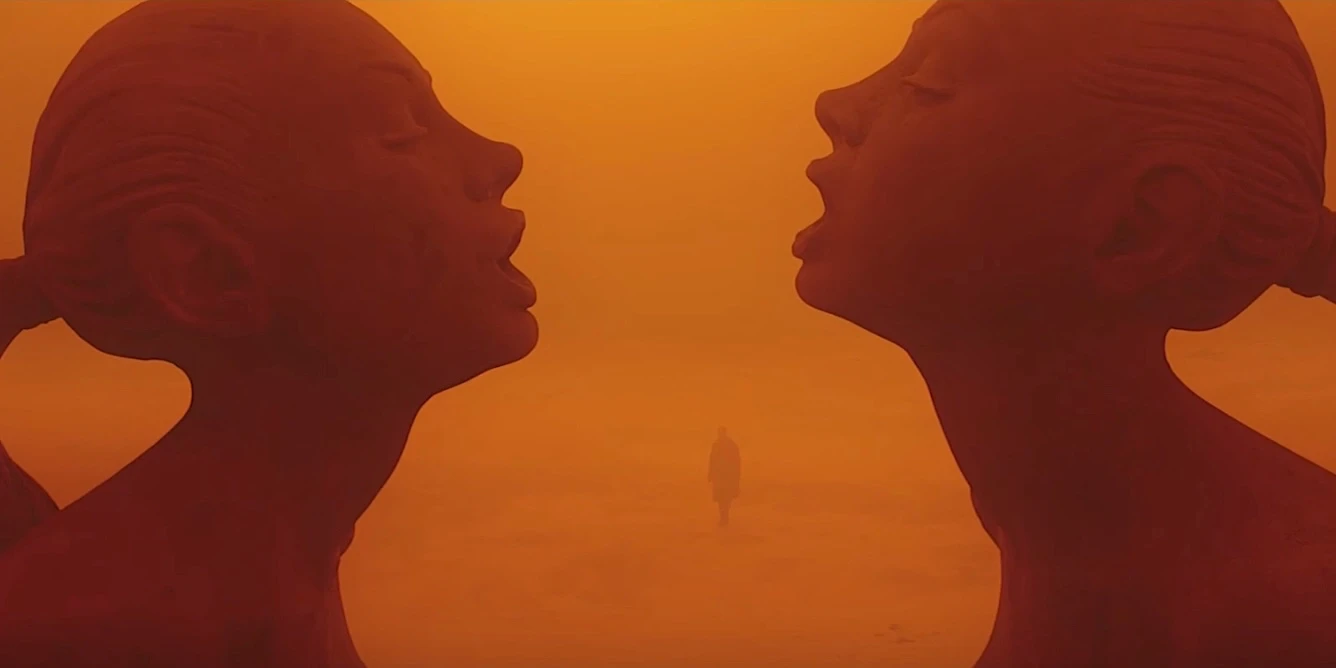 Two large dark sculptural heads facing each other in profile, a tiny solitary figure visible between them in a hazy orange desert, in deep orange and red