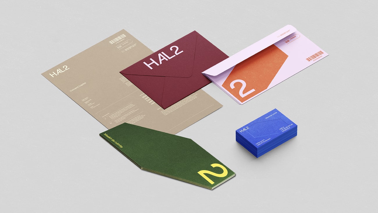 Materiais impressos exibidos em perspectiva isométrica. Folha A4 beige, envelope cor vinho, outro com rosa claro e laranja, cartão de visitas azul e outro elemento gráfico em verde escuro com fontes amarelas.