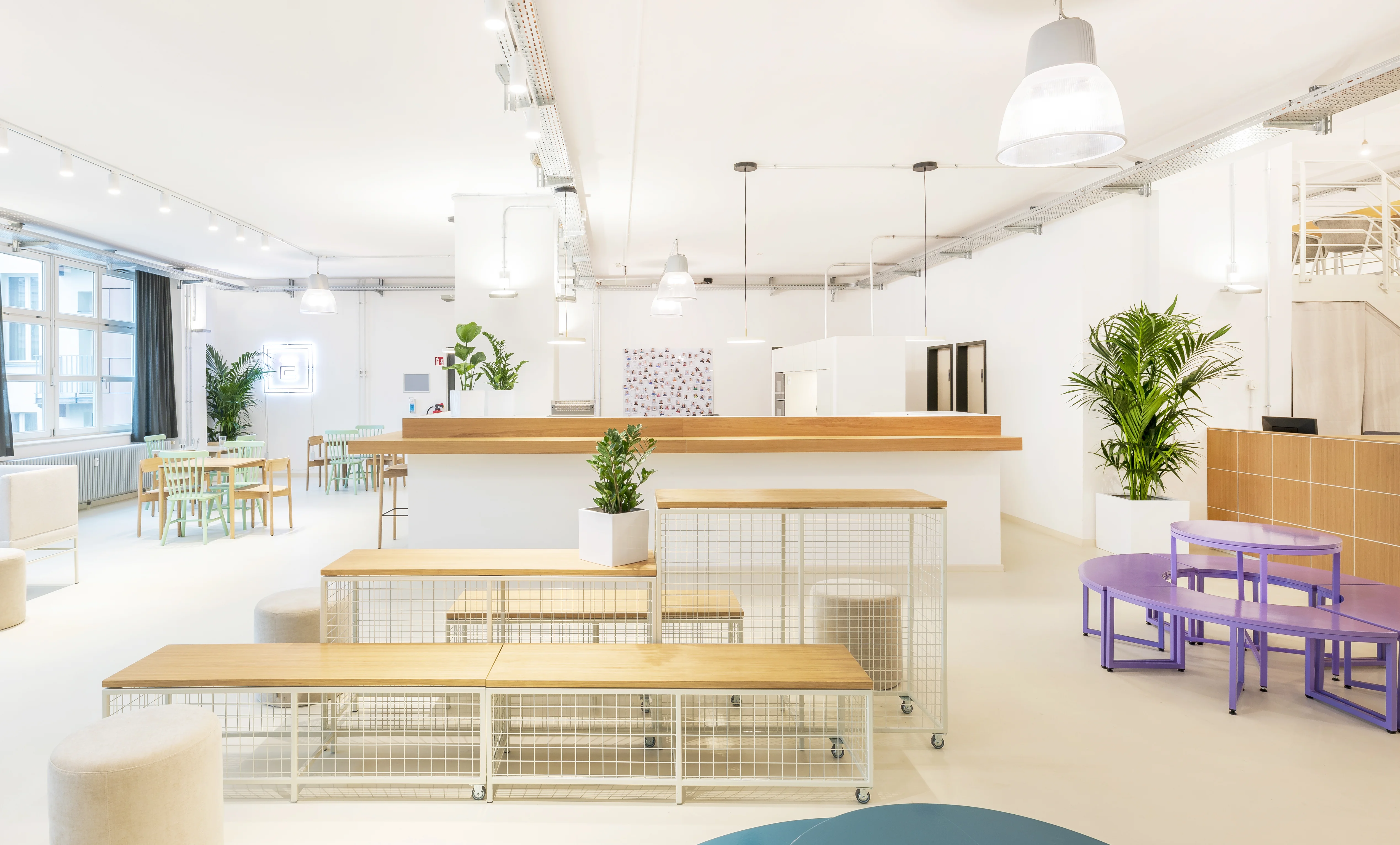 Bright open space at ELEMENT with mobile wooden benches on white mesh bases, purple curved benches and tables, a kitchen counter with oak top, mint chairs and potted palms.