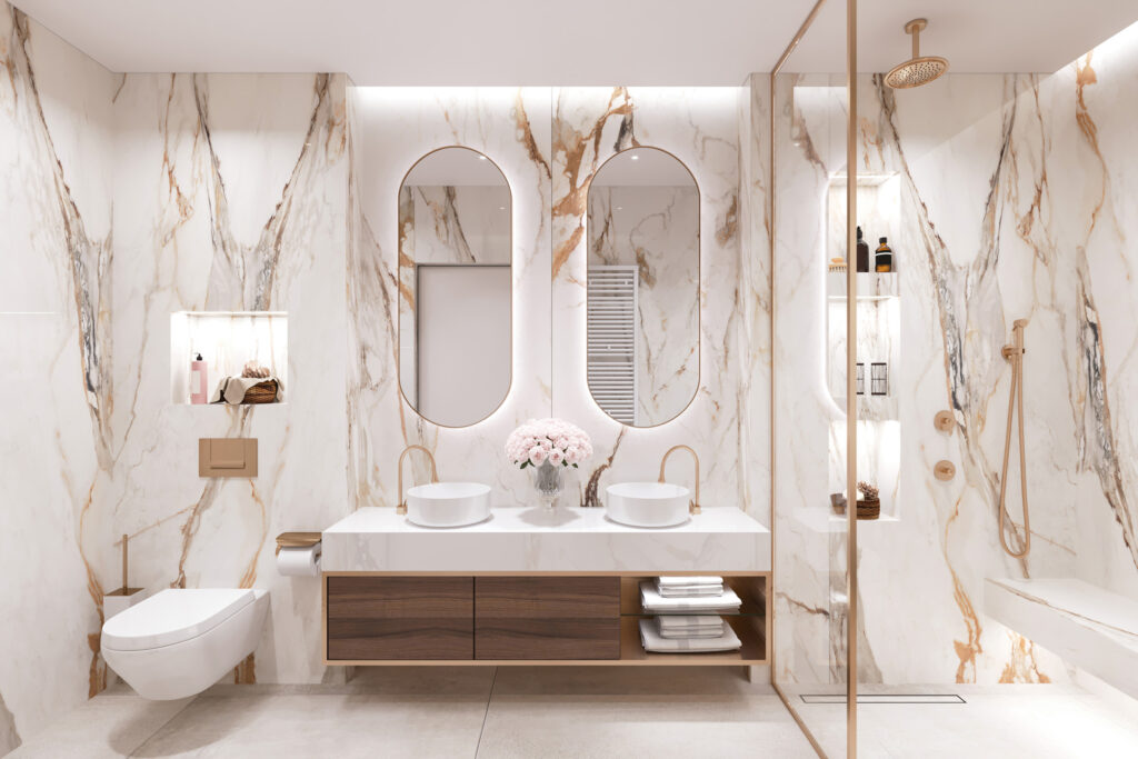 Modern bathroom with marble walls and countertops, two oval mirrors, dual white vessel sinks with gold faucets, and a vase of pink roses centered between the sinks