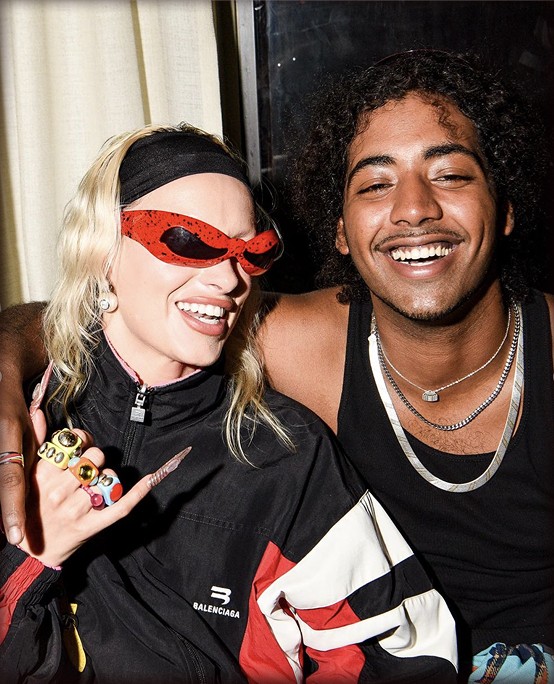 Two people posing together, one with sunglasses and a stylish outfit, both smiling and showing a playful gesture.