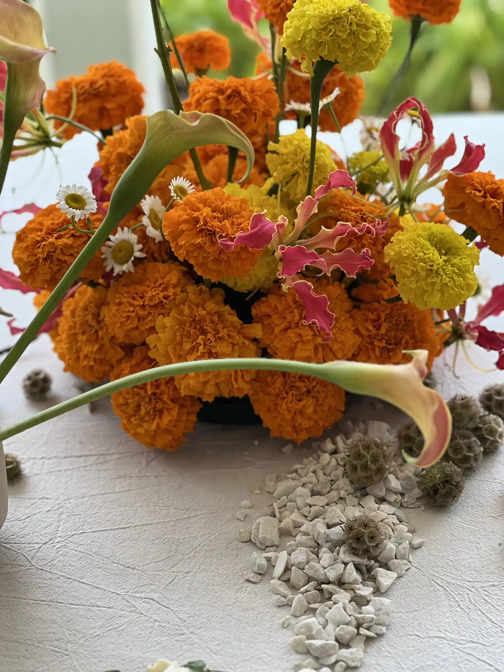 Arreglo floral colorido con calas, claveles naranjas y amarillos, margaritas pequeñas y flores exóticas rosadas, acompañado de piedras blancas decorativas sobre una mesa.
