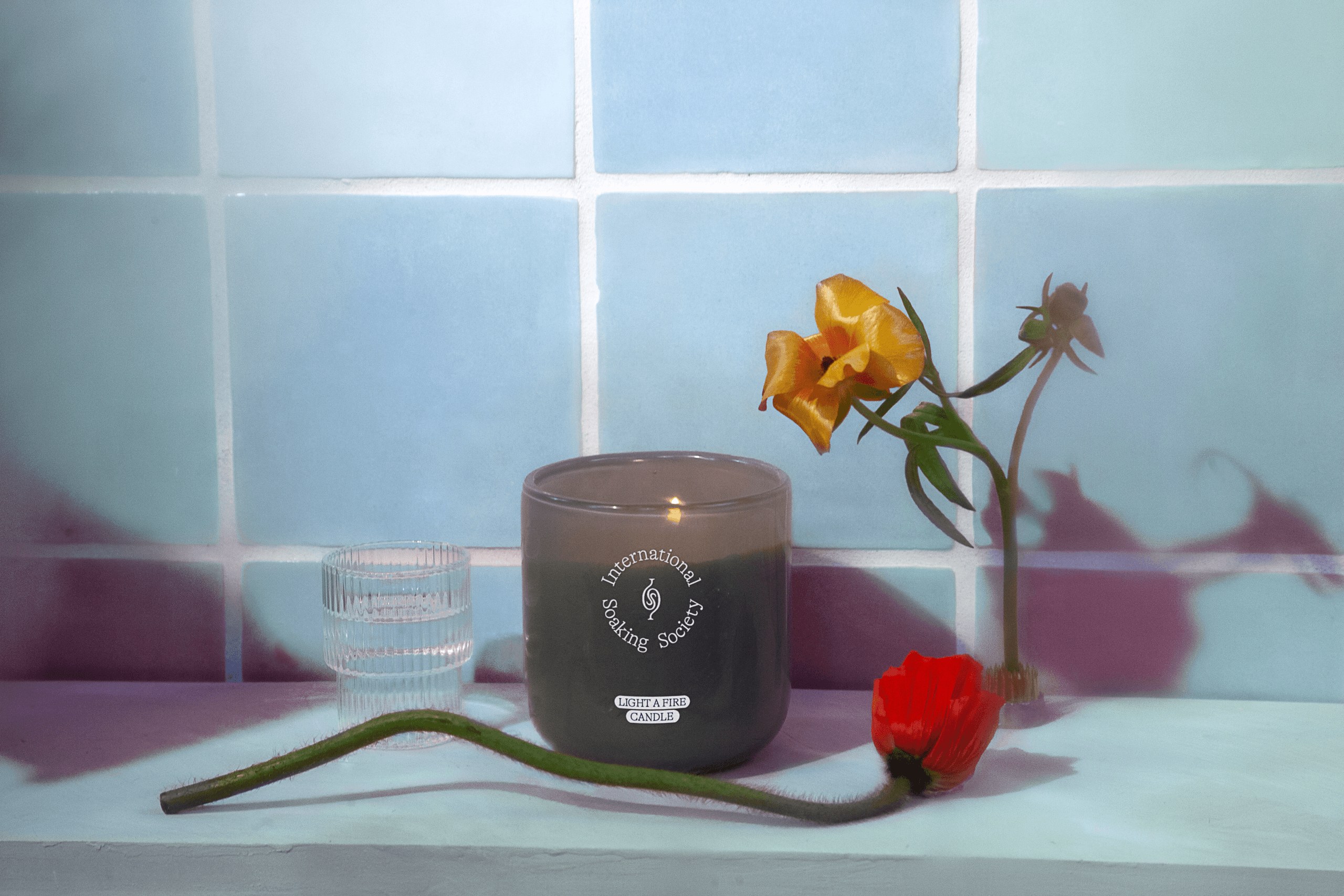 A dark gray candle in a clear glass jar sits on a light teal surface. The jar has a circular logo reading “International Scented Society”. A single orange flower with a green stem rests beside the candle. A clear glass with vertical ridges is to the left of the candle. A single red poppy lies on the surface in front of the candle. The background consists of pale blue-green square tiles.