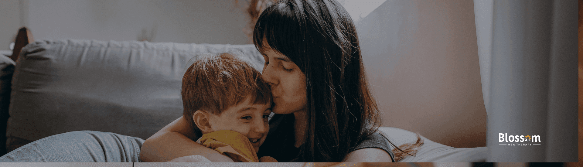 Mother warmly hugging her young child on a couch, giving him a gentle kiss as he smiles and leans into her embrace.