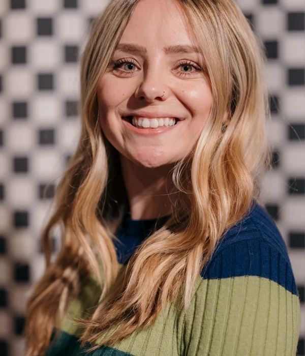 A person with long wavy blonde hair is shown from the shoulders up, wearing a striped blue and green sweater. The background features a black‑and‑white checkerboard pattern.