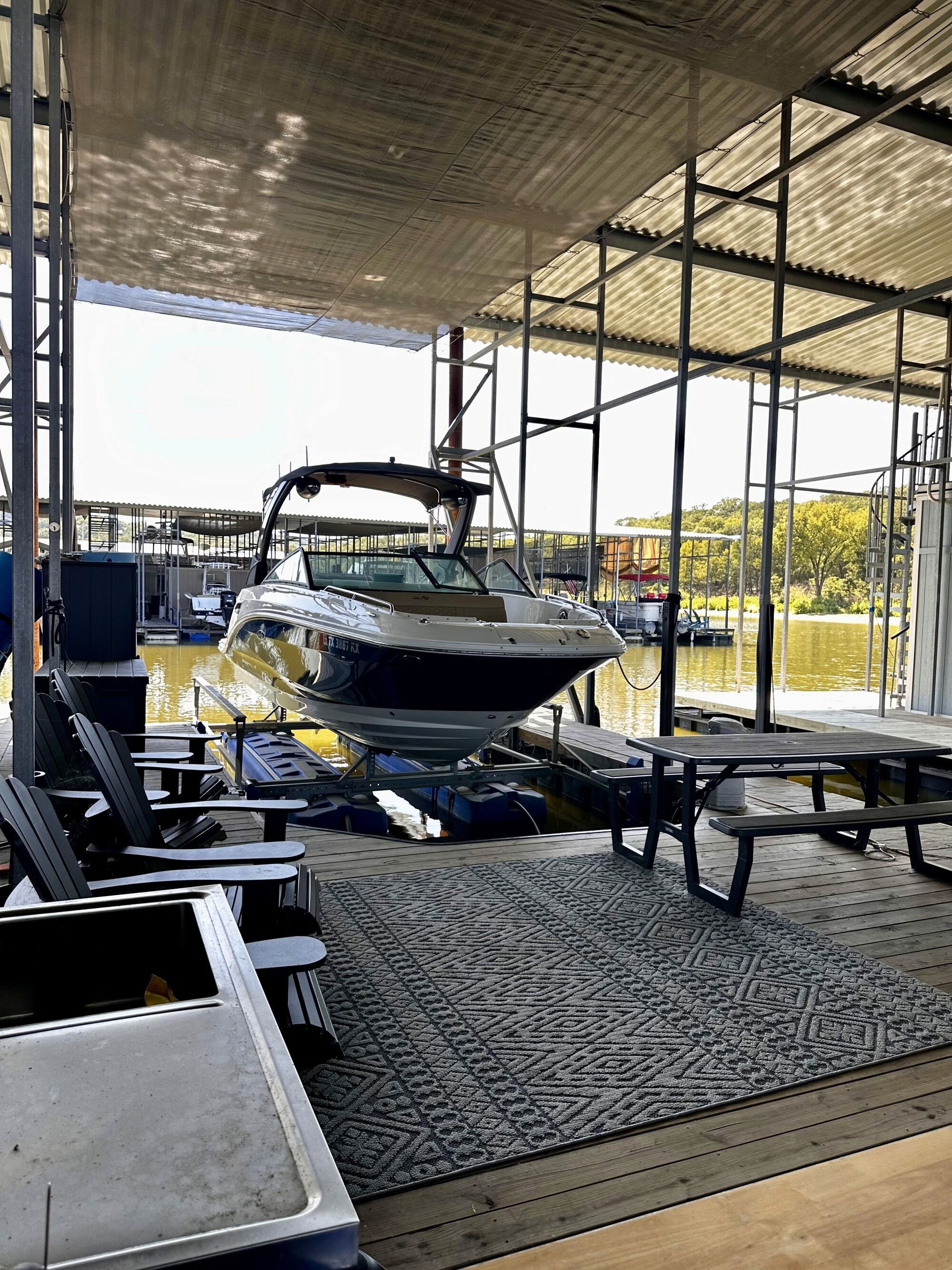 A sleek speedboat is docked on a covered marina slip, surrounded by outdoor seating and tables, with a sunlit view of the water beyond, emphasizing leisure and water activities.