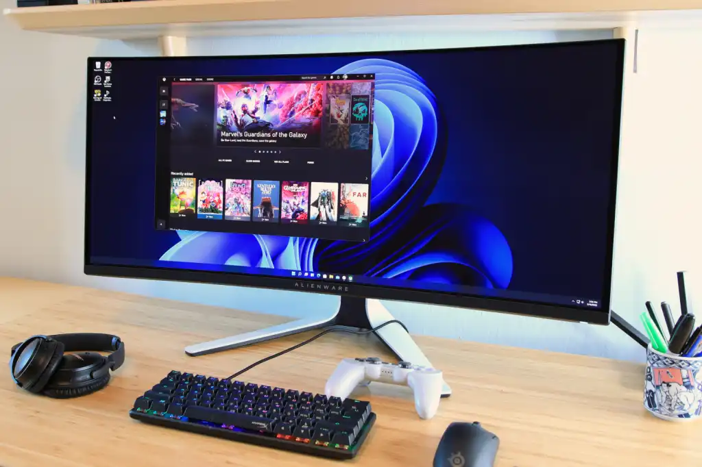 A widescreen computer monitor on a wooden desk, displaying multiple open applications, with a keyboard, mouse, and gaming controller nearby.