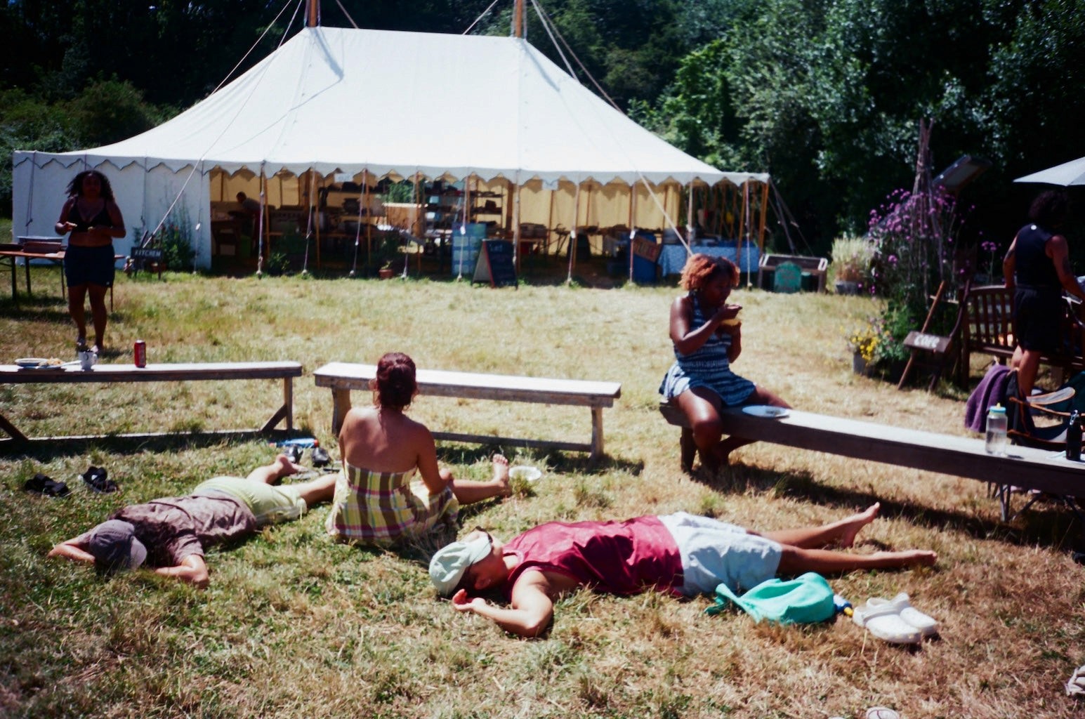 A few people sit and lie on the grass in the sun, near a festival tent
