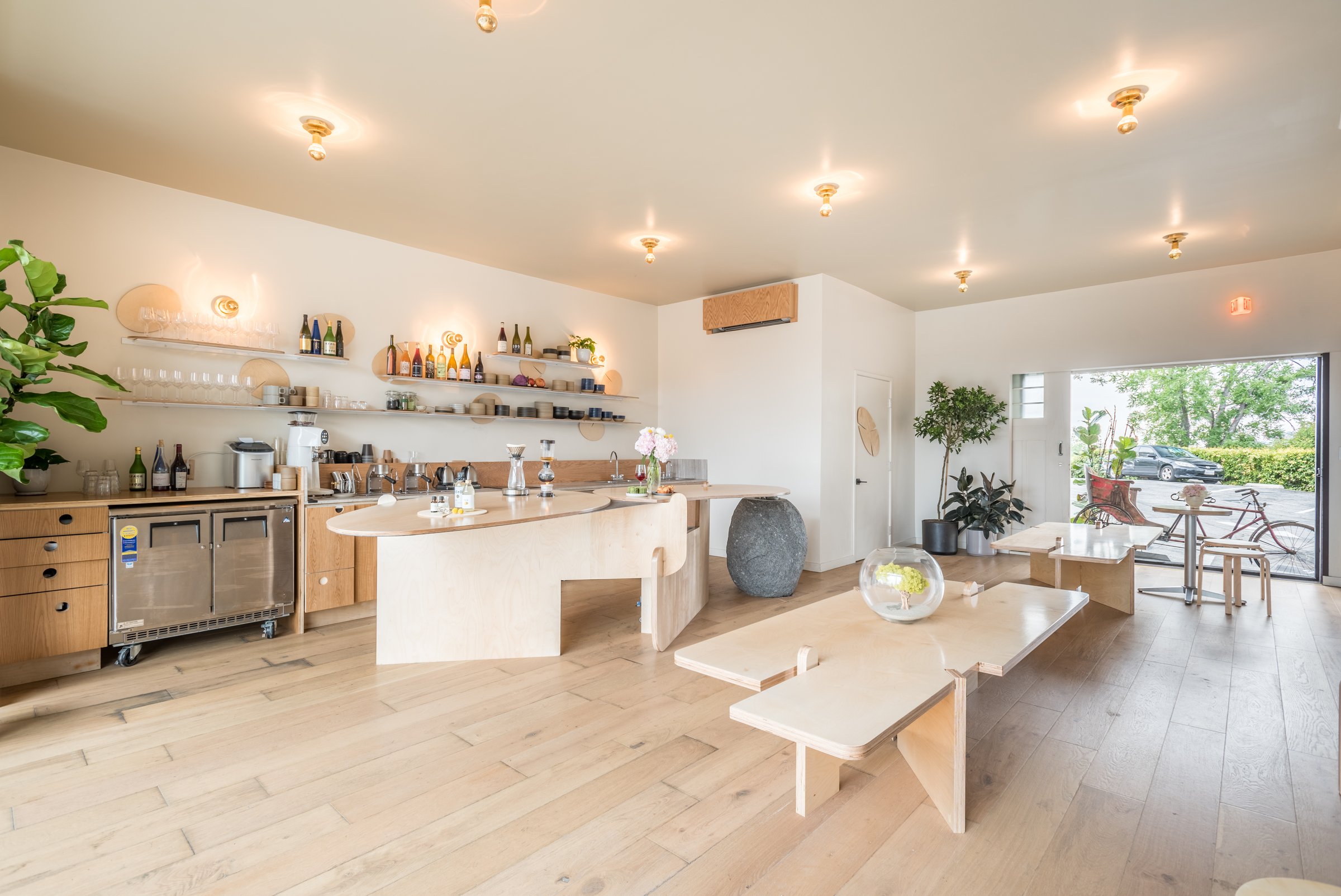 Bright dining room with a curved pale timber bar, open bottle shelves and long communal tables.