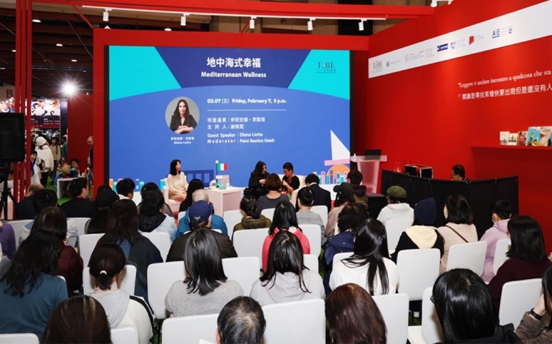 A young person with curly hair smiles at the camera while sitting in front of a laptop against a dark background.