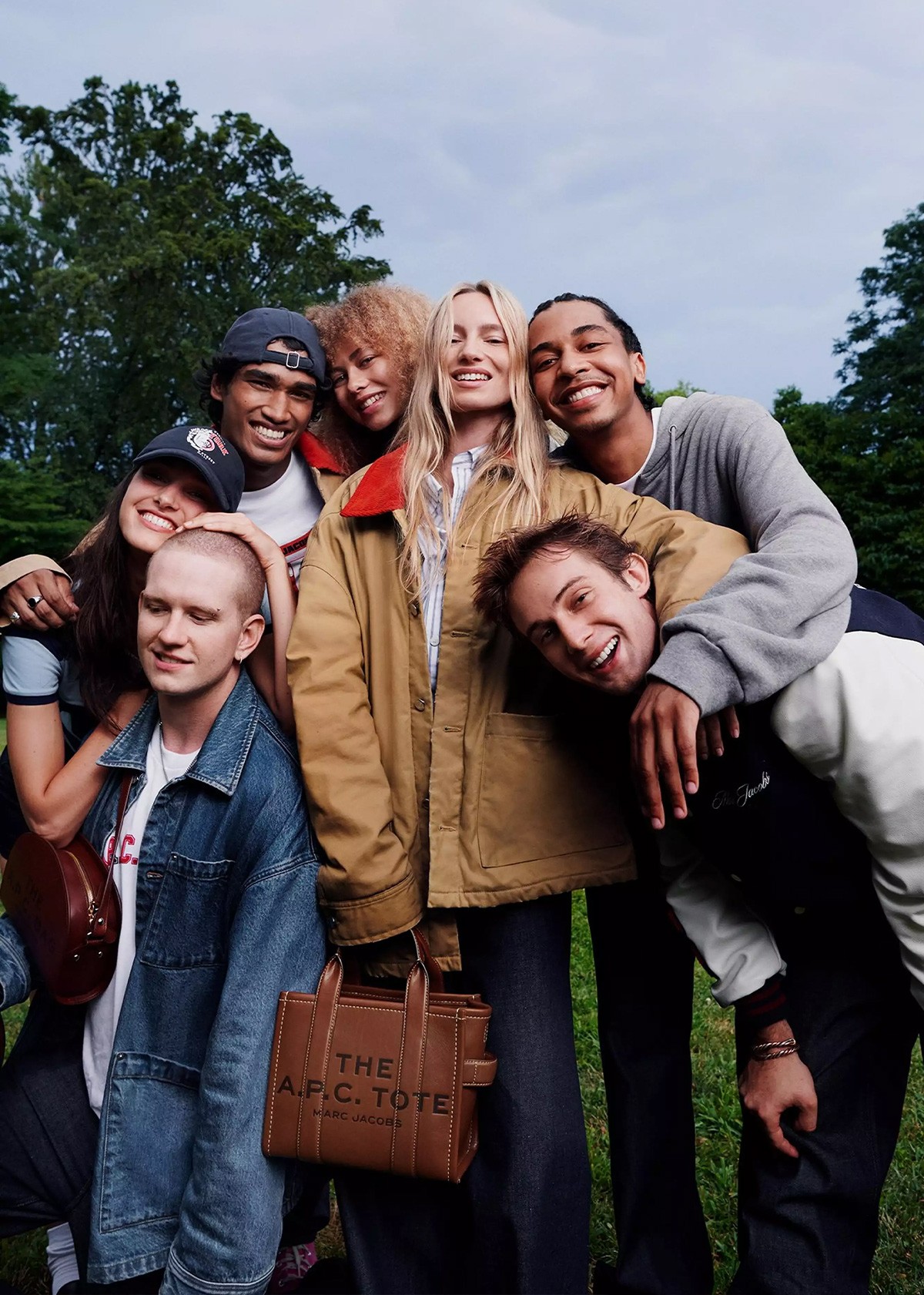 A diverse group of friends stand closely together outdoors, smiling and wearing casual jackets, with one holding a Marc Jacobs tote bag in a park setting.