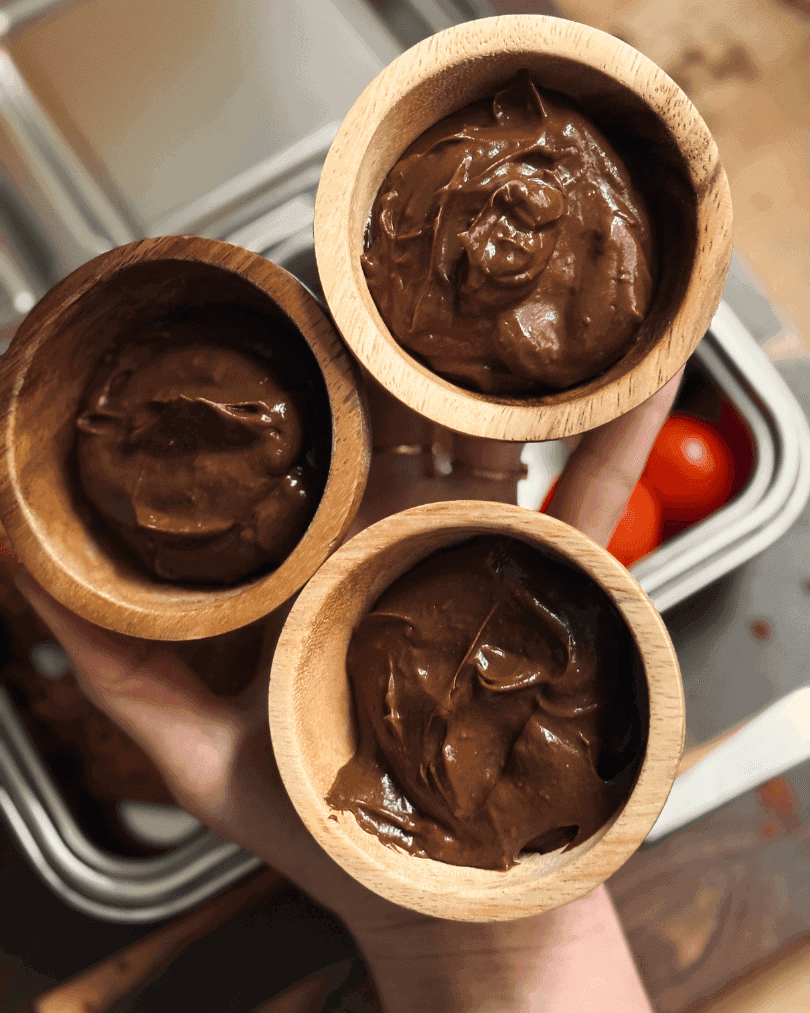 Chocolate avocado mousse served in small wooden bowls, held in a hand with lunchbox containers in the background.