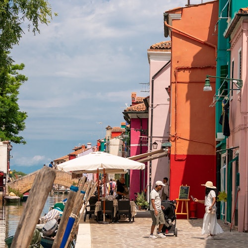 阳光明媚的环境中,运河旁一条色彩缤纷的街道,两侧是充满活力的建筑、户外座位区,还有行人悠闲漫步。