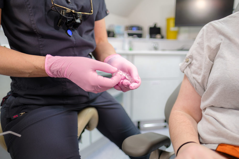 A close-up of a dental professional's hands wearing pink gloves, holding a clear Invisalign aligner to show a patient during a consultation. The dentist is wearing dark grey scrubs with dental loupes around their neck.