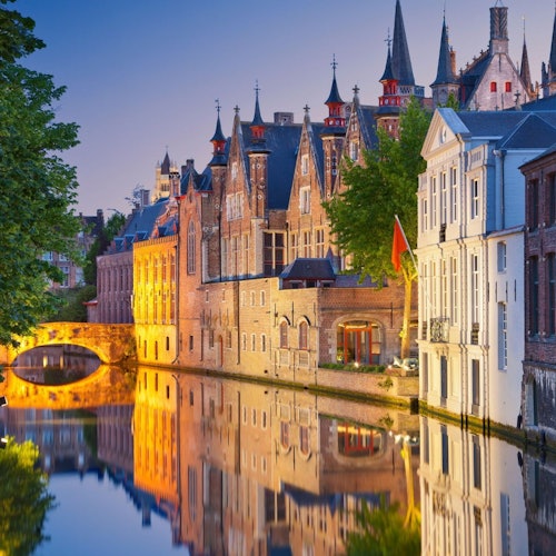 Povijesne zgrade koje se ogledaju u mirnom kanalu u sumrak, s raskošnom arhitekturom i kamenim mostom u pozadini.