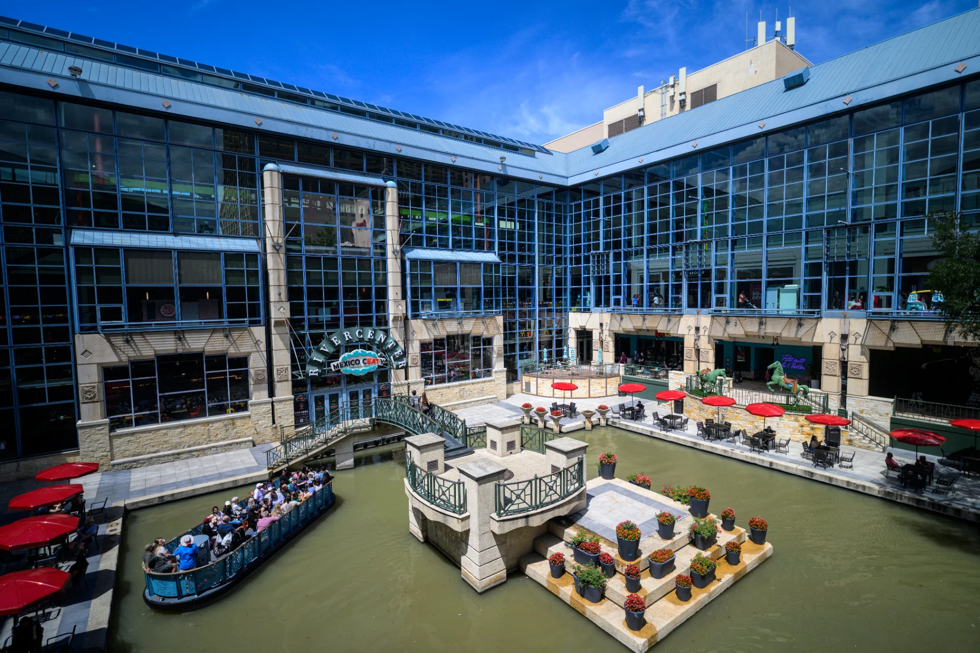 Aerial view of the Rivercenter courtyard on the San Antonio River Walk, with a river barge passing the Mexico Ceaty entrance and red-umbrella dining along the water