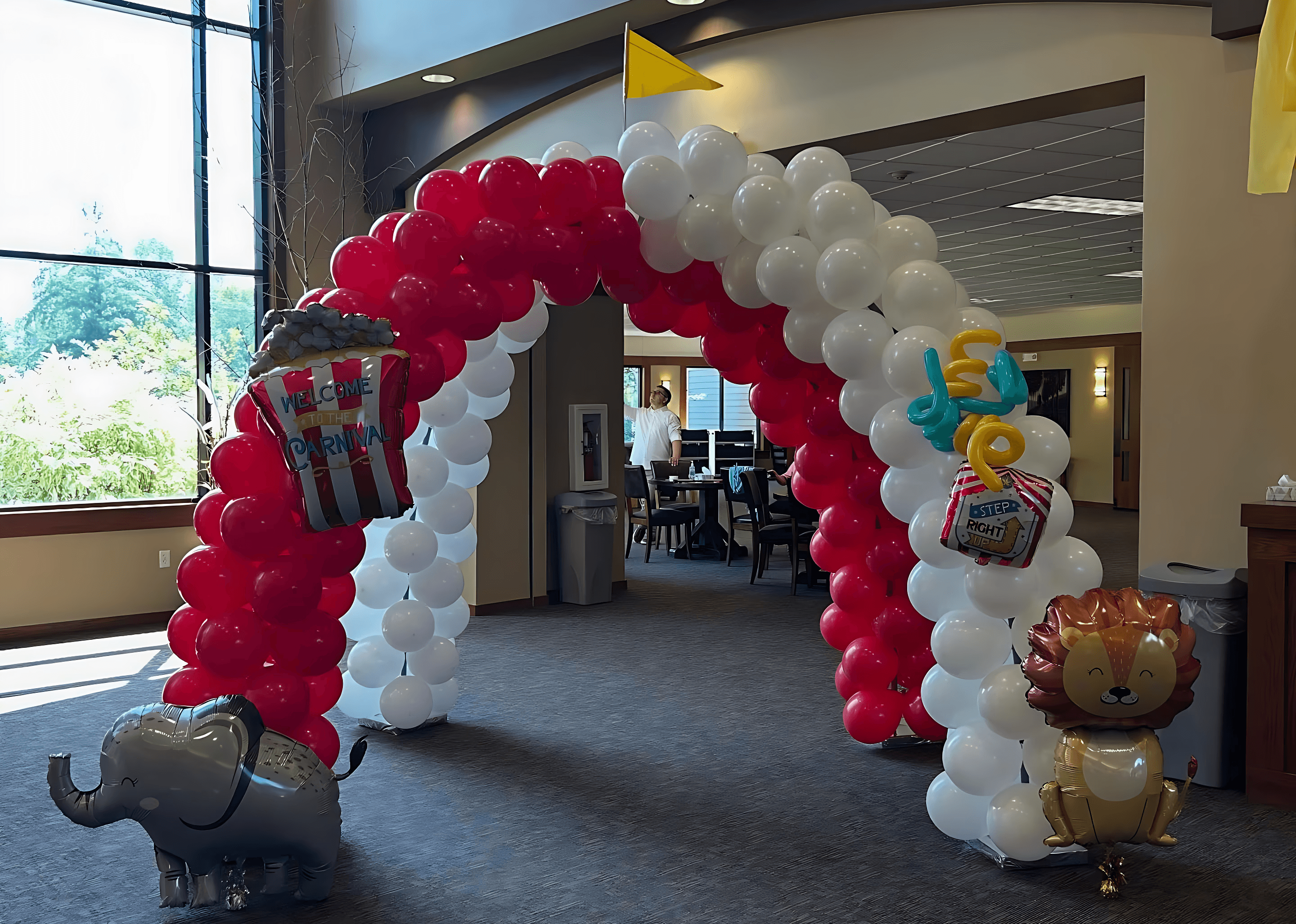 A festive red and white balloon arch, adorned with popcorn, lion, and elephant-shaped balloons, stands in a well-lit space with large windows and a glimpse of people in the background, suggesting a playful, circus-themed event.