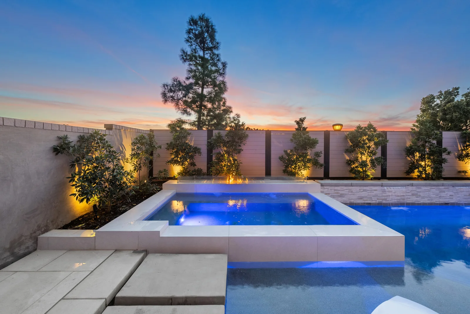 Pool and spa at sunset Illuminated pool and raised spa with pale coping, poolside steps and a fire feature at sunset.