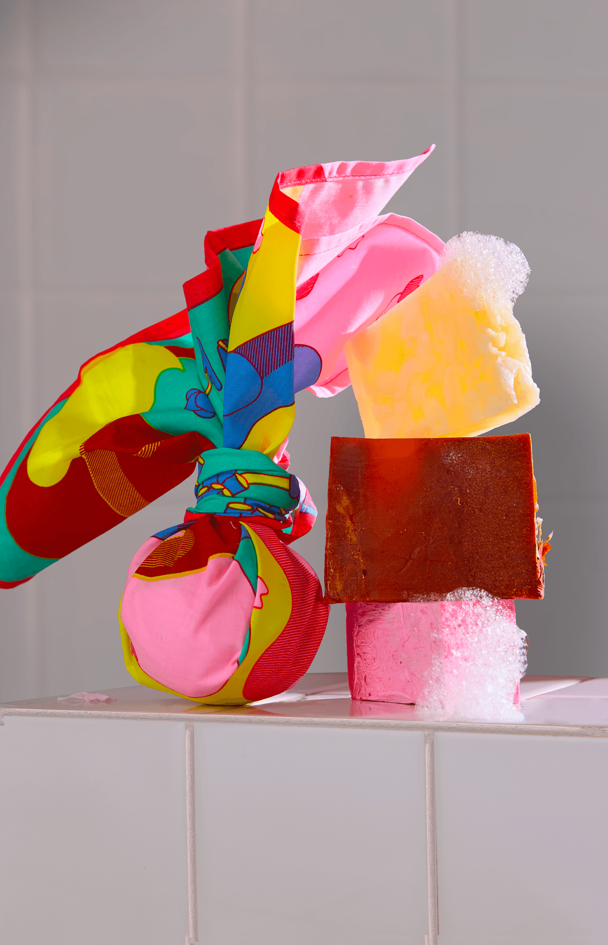 Knot-wrapped bath bomb using a colorful illustrated fabric, displayed with stacked bath products on a tiled surface.