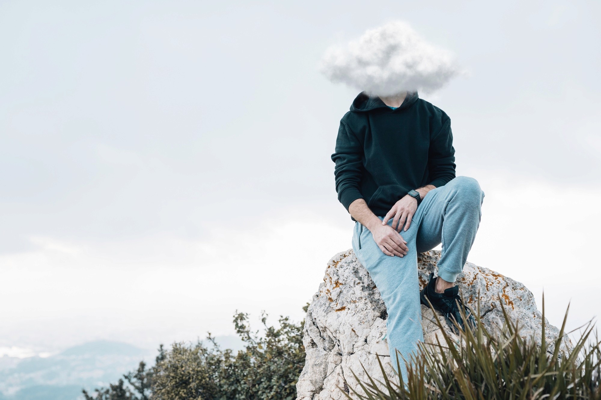 A person sitting on a rock, wearing a white cloud-shaped headpiece against a cloudy sky.