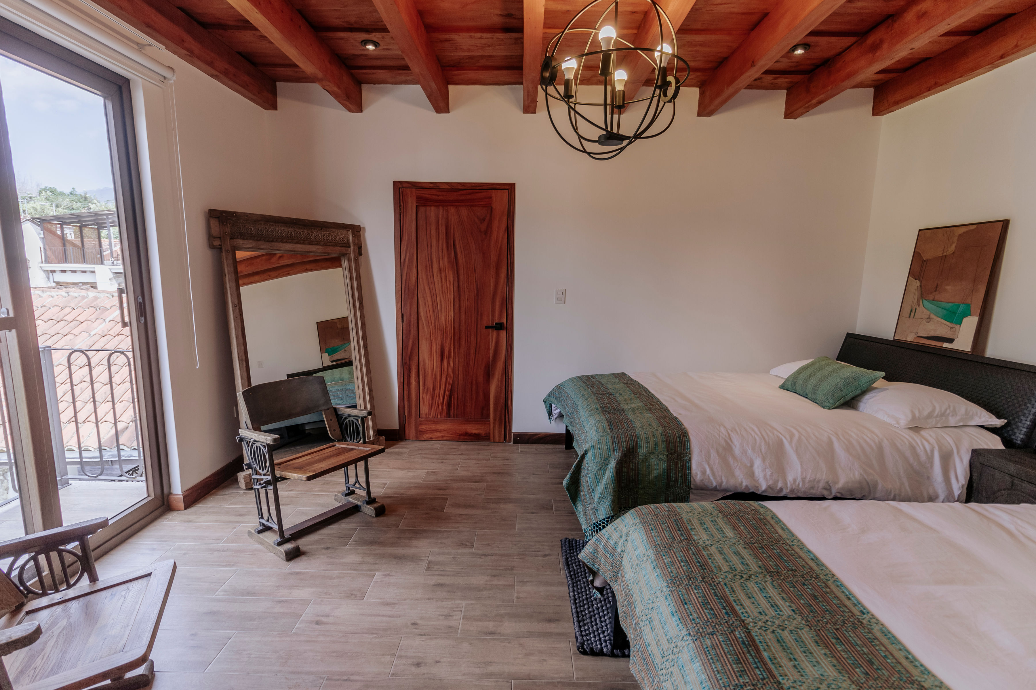 The image shows a cozy bedroom with wooden ceiling beams, a chandelier, two neatly made beds with green and white linens, a wooden door, a large mirror resting on the floor, and a balcony with a view outside, creating a warm and inviting atmosphere.