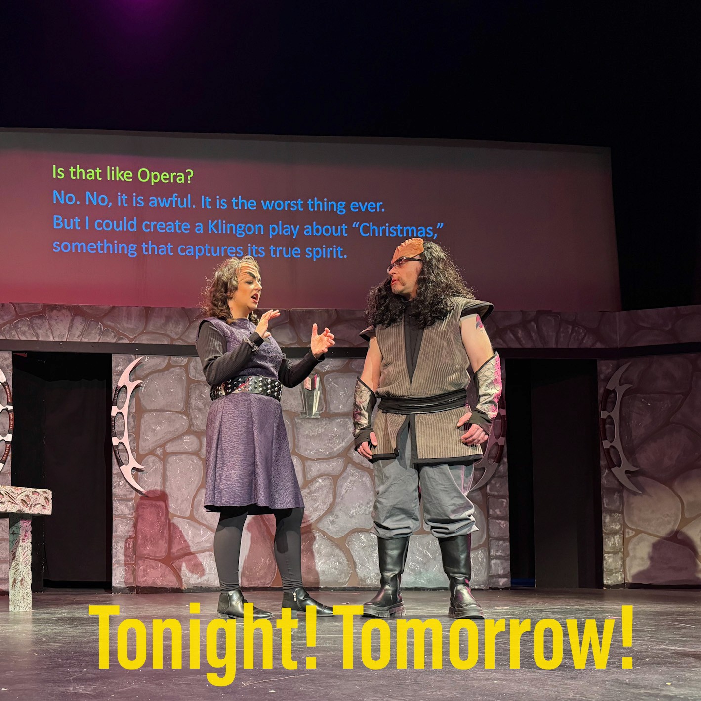 Two theater actors performing a dramatic scene on a stage in front of a painted stone wall backdrop.