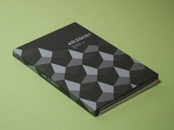 A book with a black cover featuring a geometric pattern of gray and white triangles rests on a light green surface. The title "ARLEQUIN" is printed in white on the cover, and the spine also displays "ARLEQUIN" in white. The book is positioned diagonally, with its cover facing the viewer.
