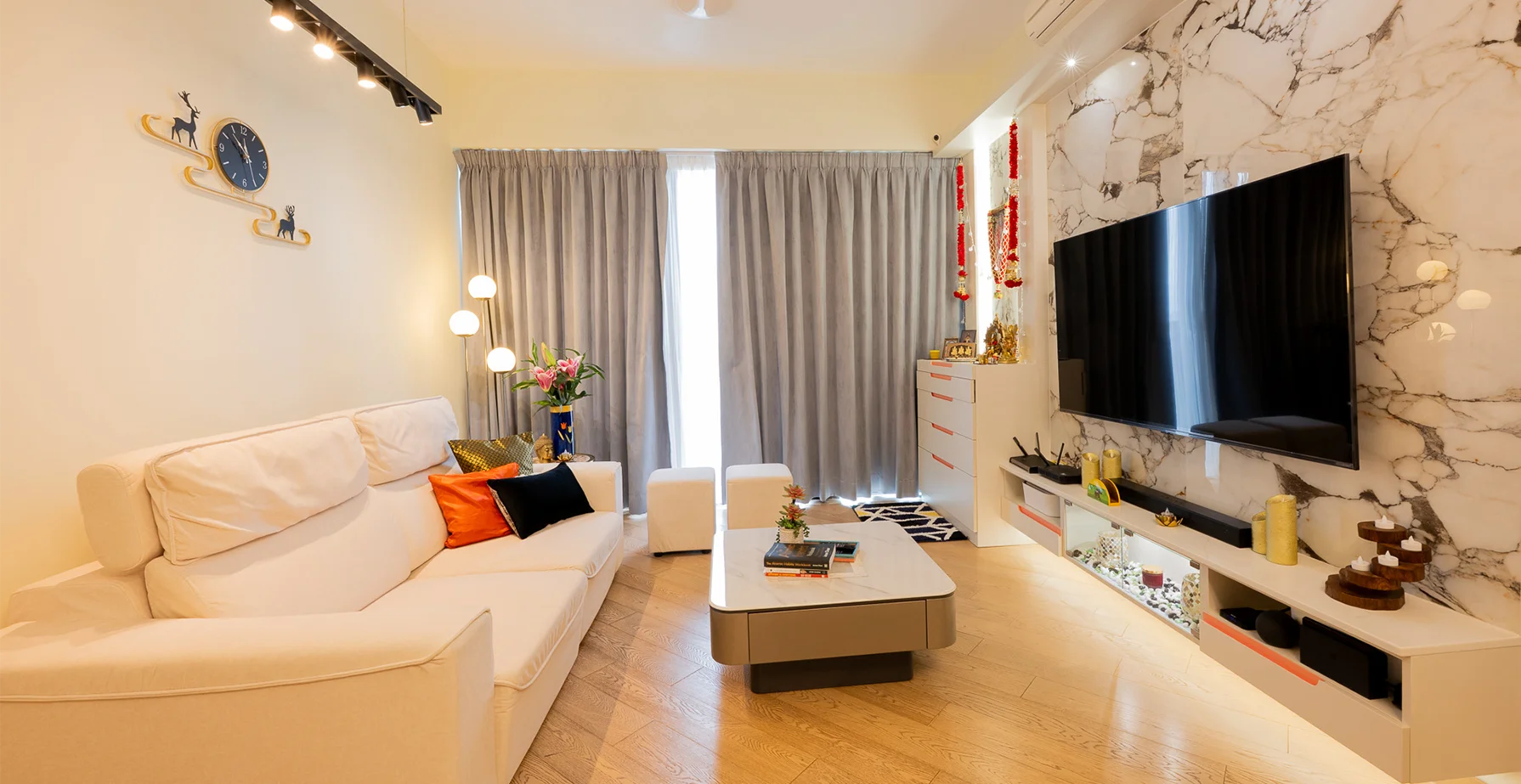 Living room with a white sectional sofa, orange and black cushions, marble-effect TV wall, and built-in media storage.