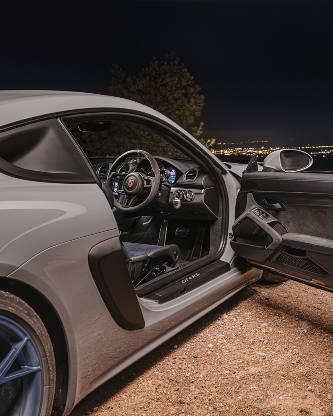 Grey Porsche 718 Cayman GT4 RS with the driver's door open, revealing the Alcantara steering wheel, carbon bucket seat and "GT4 RS" door-sill plate, with distant city lights across the valley at night.