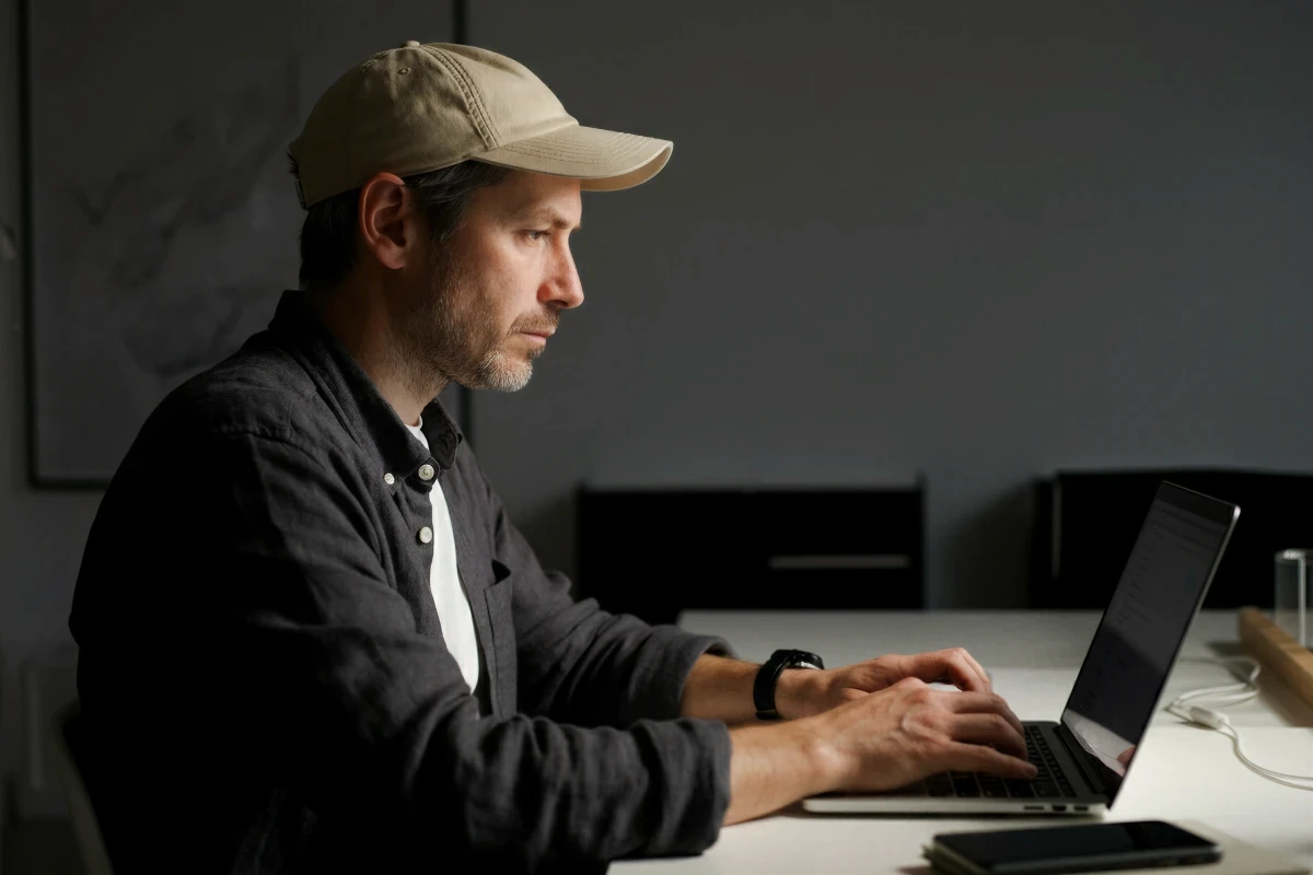 A person with a cap is focused on typing on a laptop in a minimalistic, well-lit workspace.
