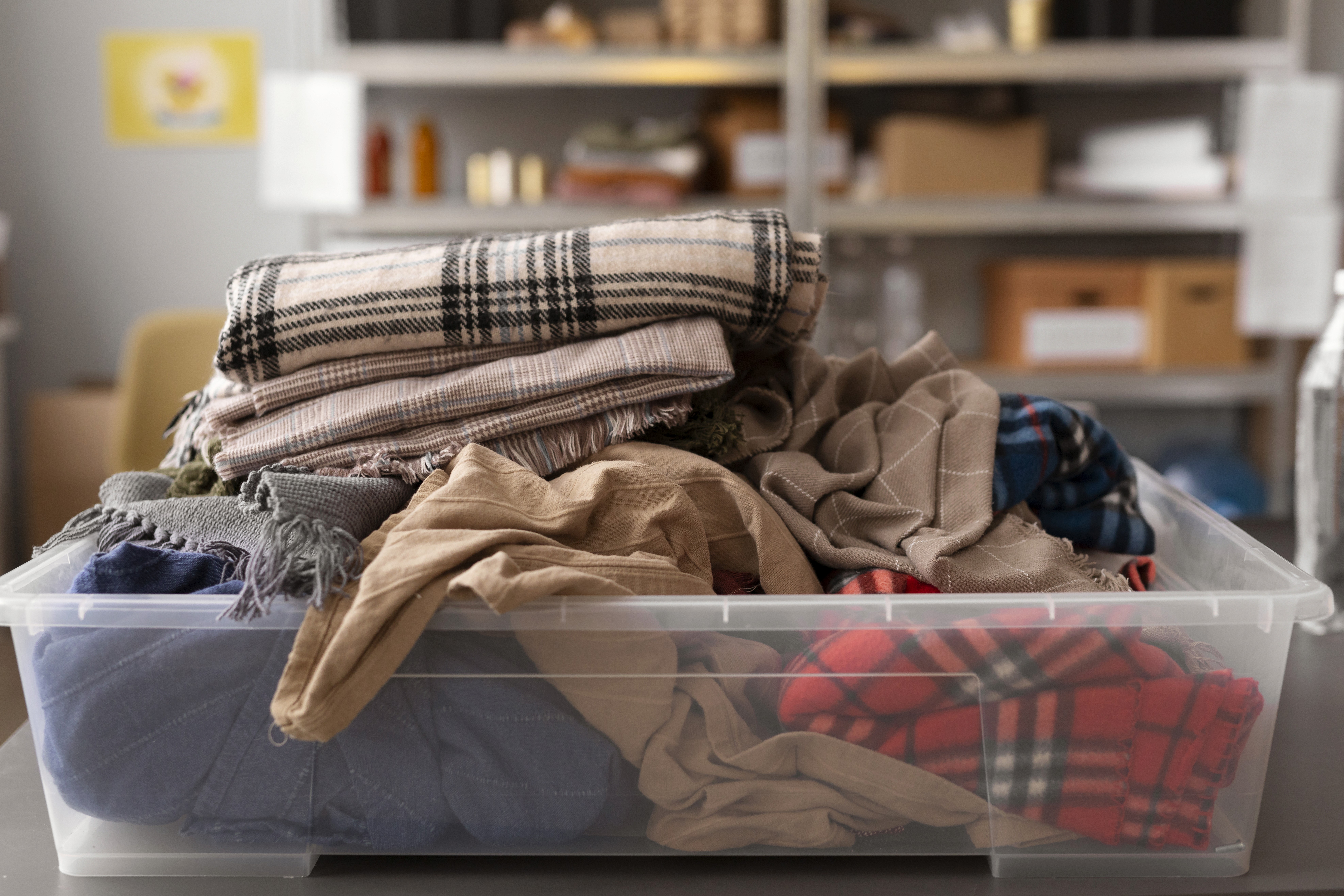 A clear plastic bin filled with various folded scarves, including plaid and solid colors, sits on a table. Shelves with boxes and items are blurred in the background.