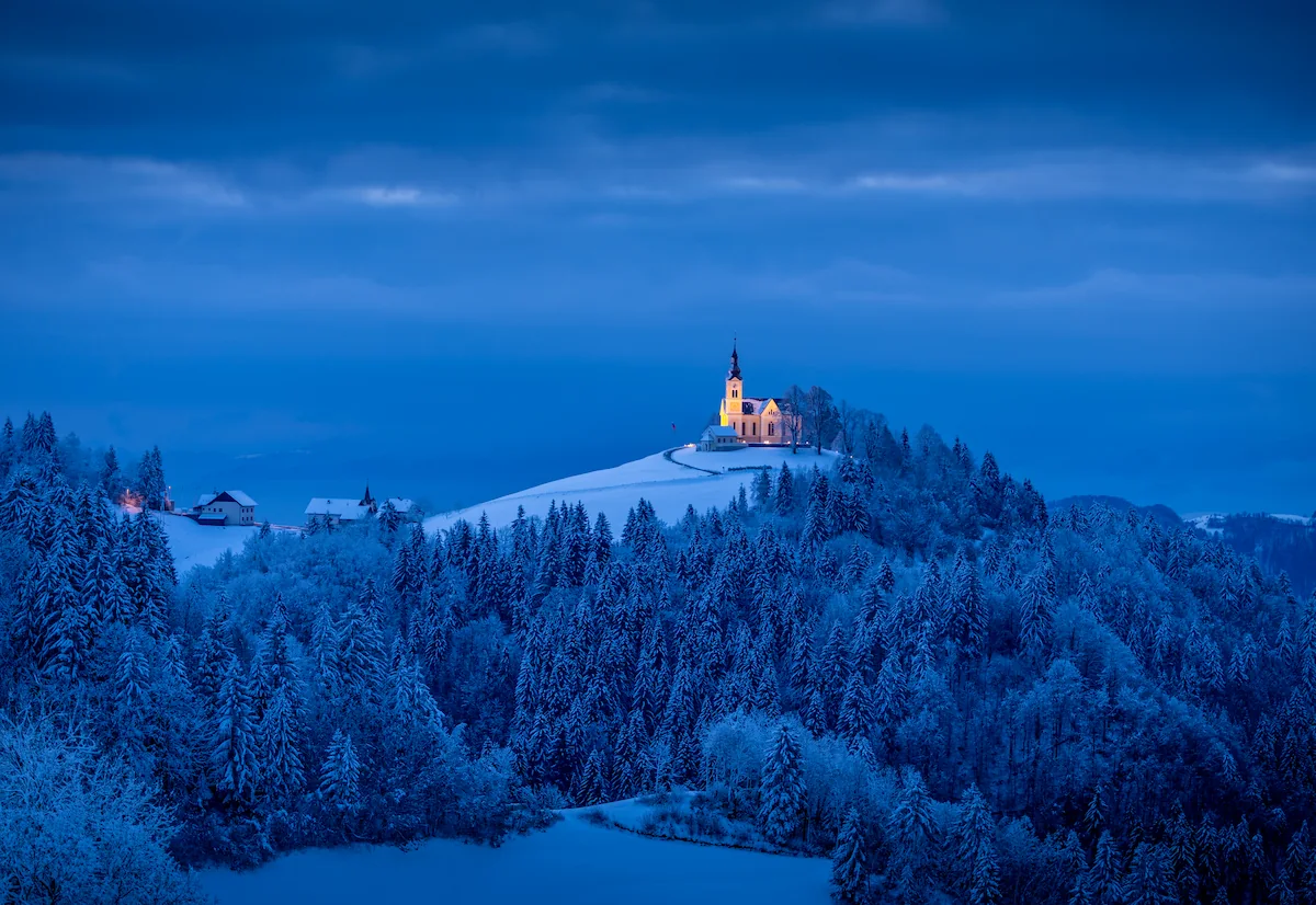 A serene blue landscape features a small church on a hill in Slovenia, surrounded by trees under a twilight sky.