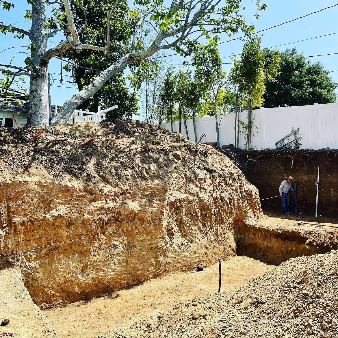 A large excavated dirt pit for a building or pool project, with a construction worker measuring the site.