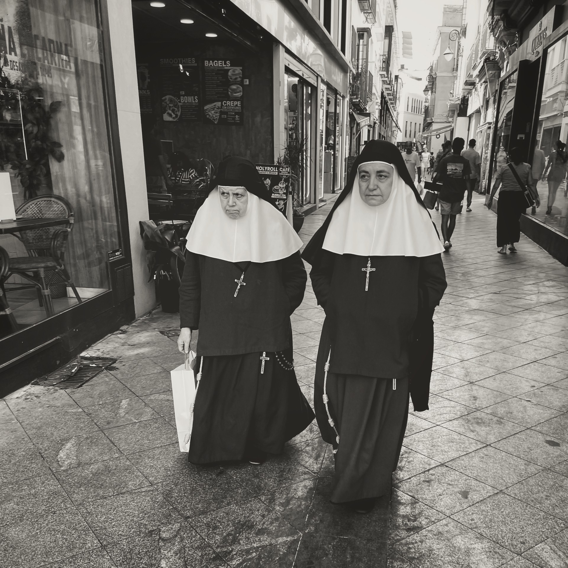 Two nuns wearing traditional habits walk down a city sidewalk surrounded by shops and pedestrians.