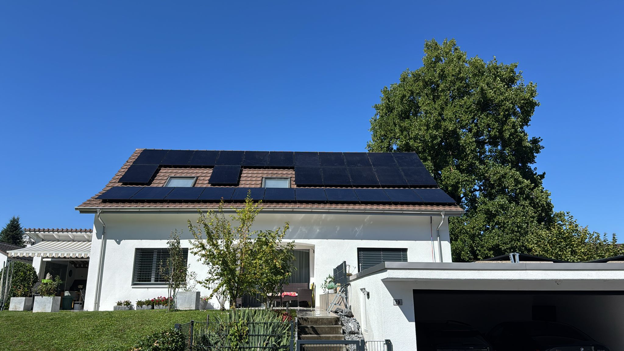 Ein weisses Wohnhaus mit einem grossen Baum im Garten. Auf dem Dach sind mehrere Reihen schwarzer Solarpaneele montiert.