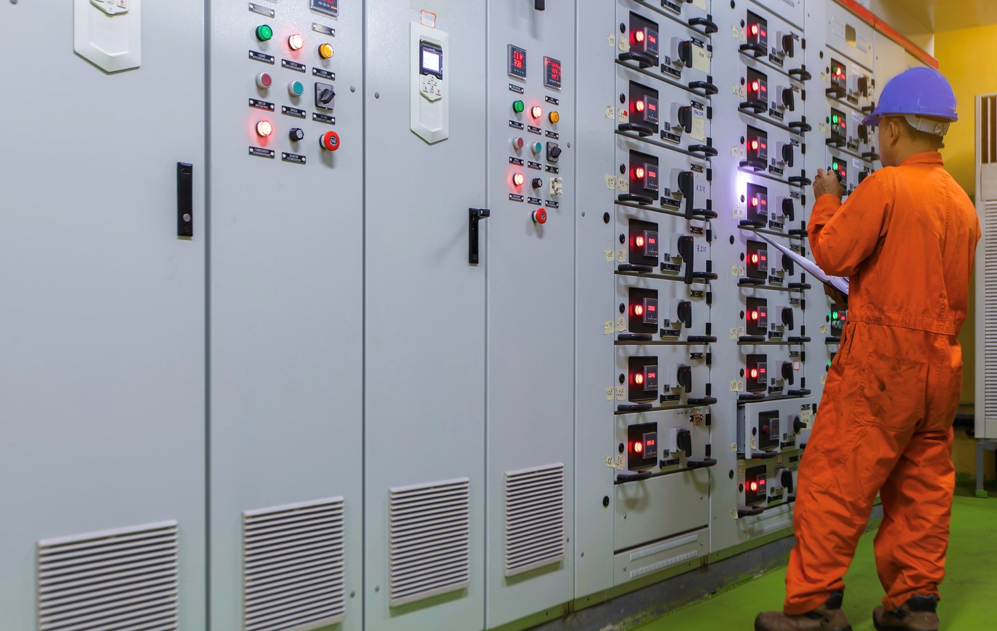 A worker in an orange jumpsuit inspects electrical control panels in a facility.