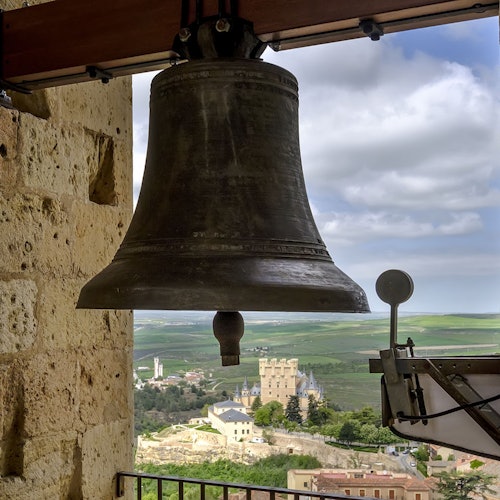 Large bronze bell in a stone tower with a view of a distant castle and countryside under a partly cloudy sky.