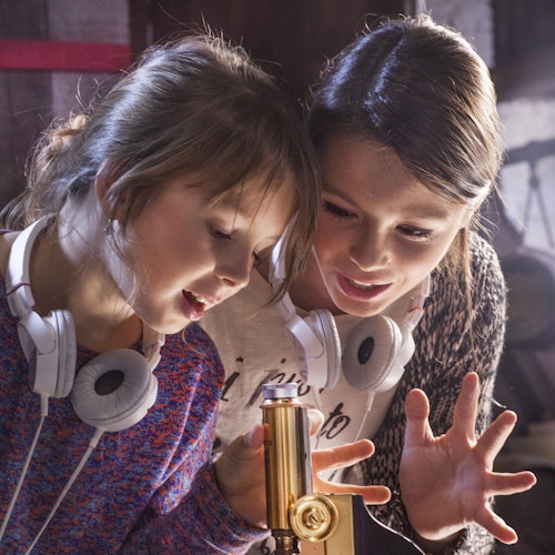 Két lány, akik nyakuk körül fejhallgatót viselnek, kíváncsian vizsgálnak egy réztárgyat, valószínűleg egy mikroszkópot, egy szobában.
