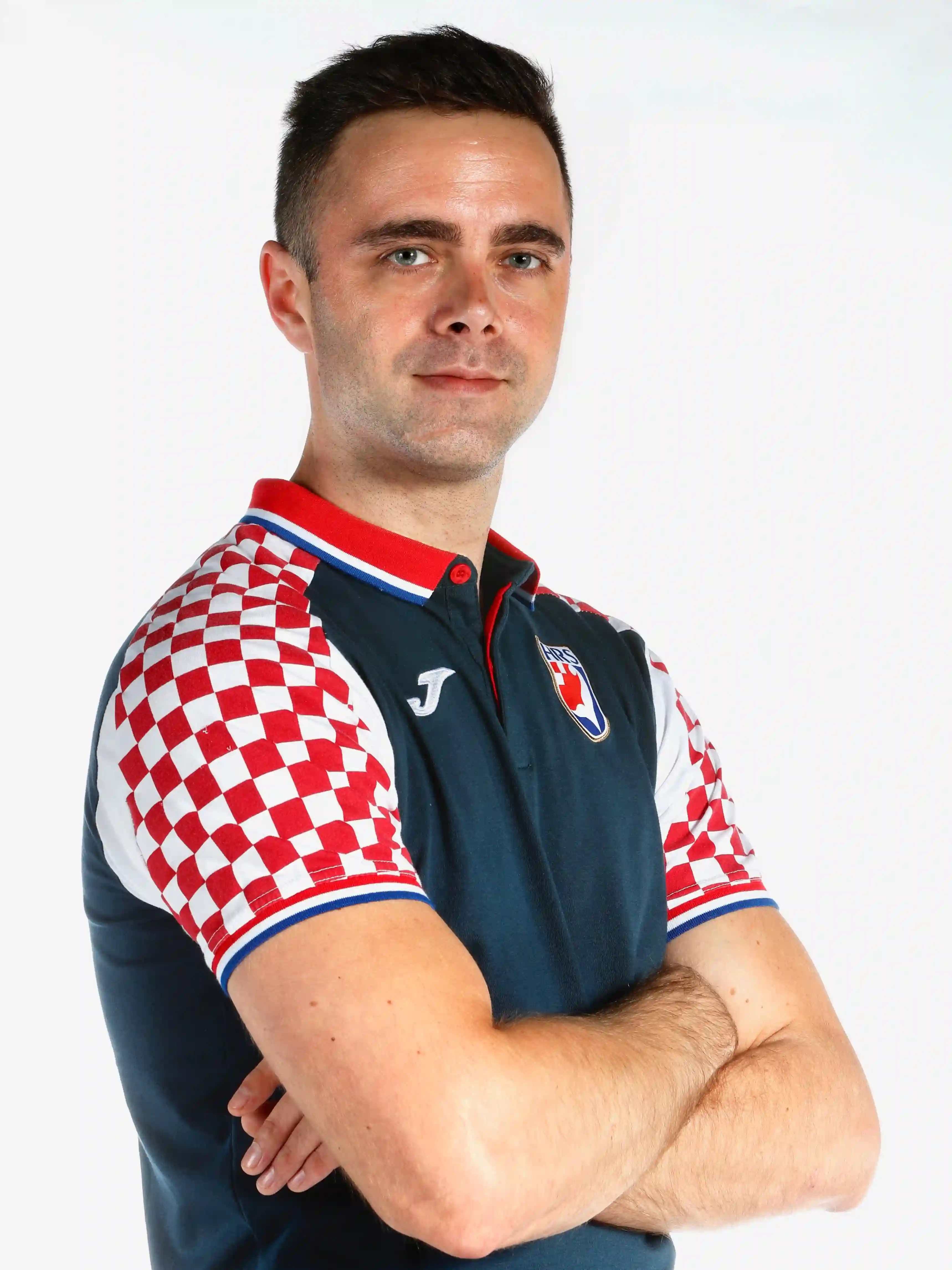 A smiling young man with crossed arms, wearing a plaid shirt and white t-shirt, poses against a dark background.