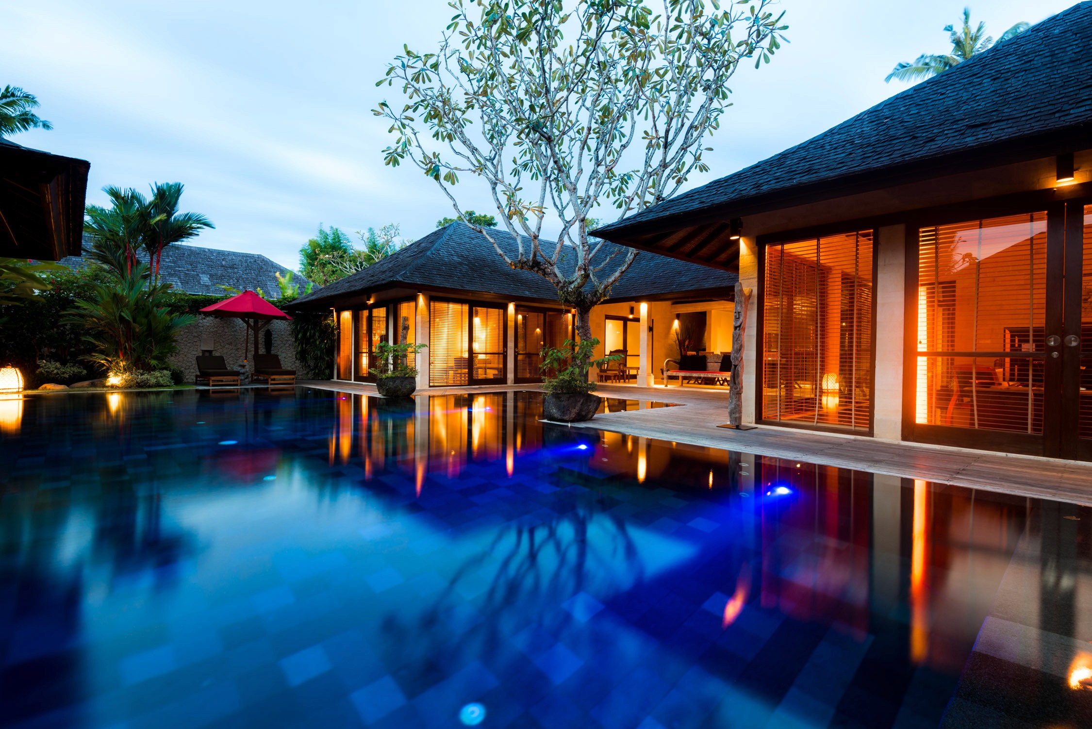 Tropical resort villas with thatched roofs and warm interior lighting reflected in a large swimming pool at dusk.