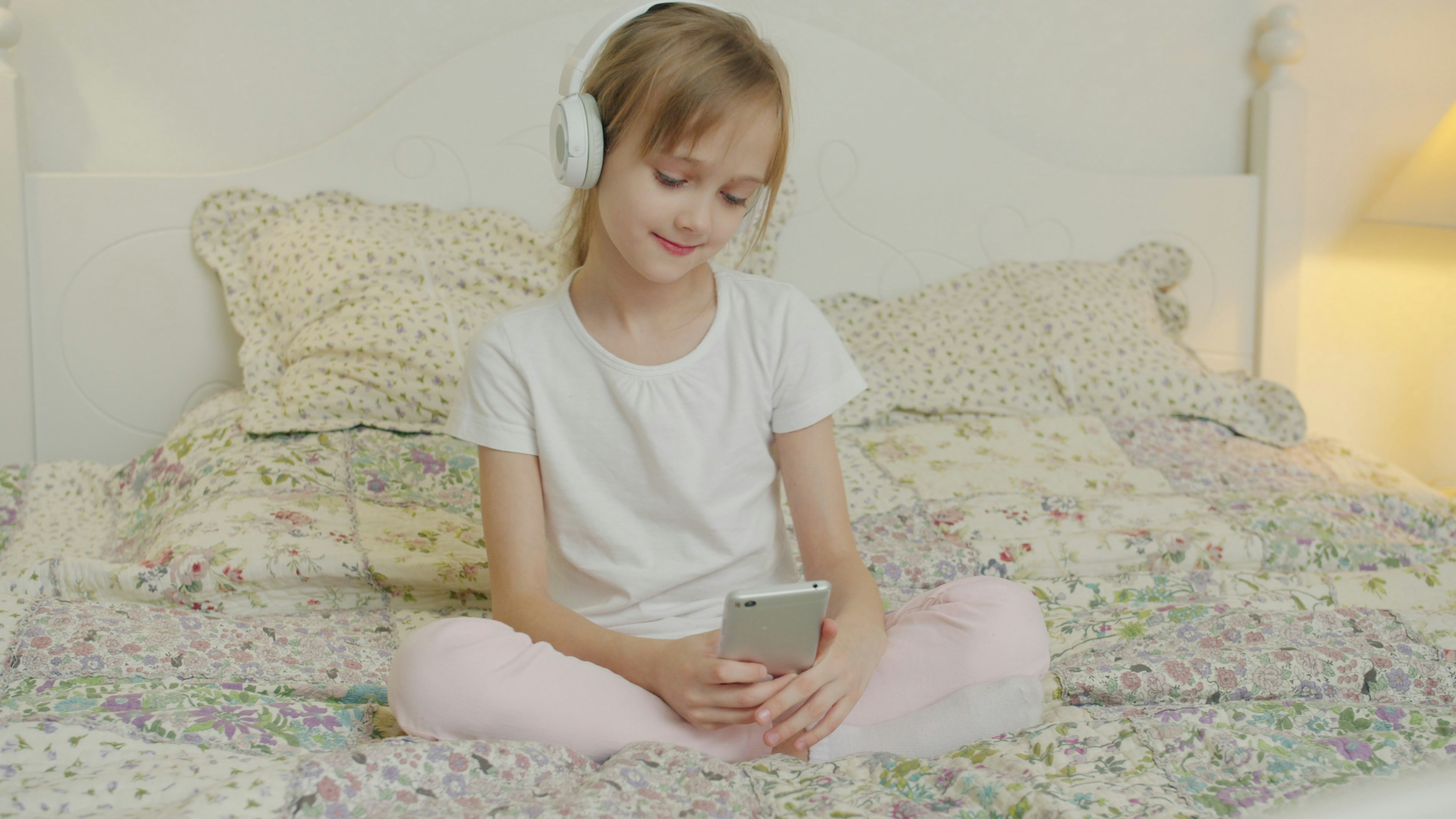 Young girl wearing headphones and using a smartphone while sitting on a bed.