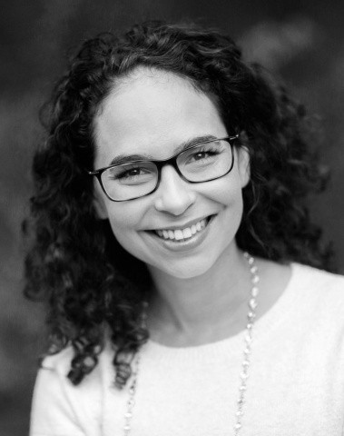 A woman with curly hair and glasses smiles at the camera, wearing a light-colored top against a blurred background.